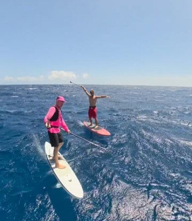 Dos riders de SUP foil en pleno downwind sobre el mar azul, uno con chaleco rosa remando y otro saludando